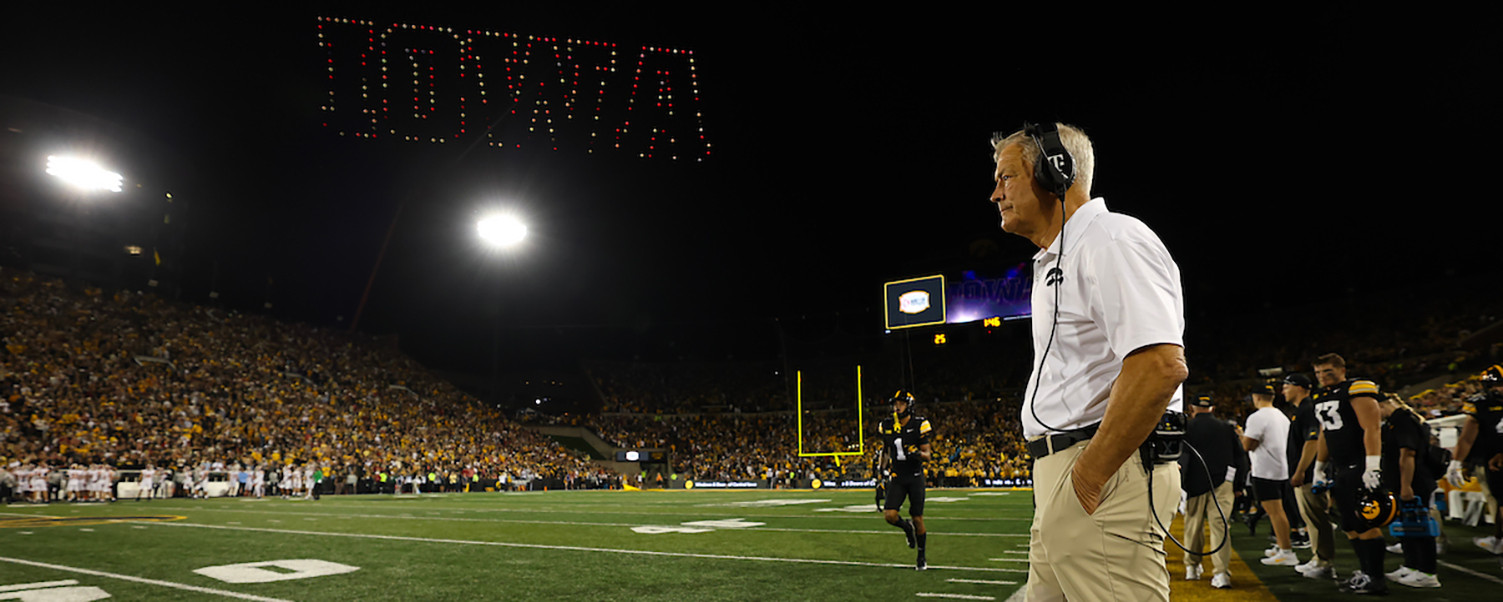 Kirk Ferentz stands on the sidelines of a football field at night, with a large "IOWA" sign illuminated overhead and a crowd in the stands.