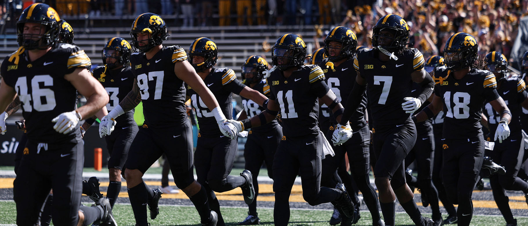 A group of American football players in black uniforms with yellow accents run onto the field, with a large crowd in the stands behind them.