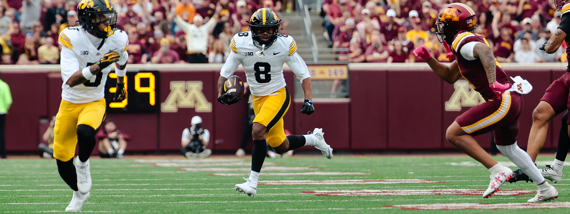 A football player in a white and yellow uniform runs with the ball while being pursued by opponents on a field with a crowd in the background.